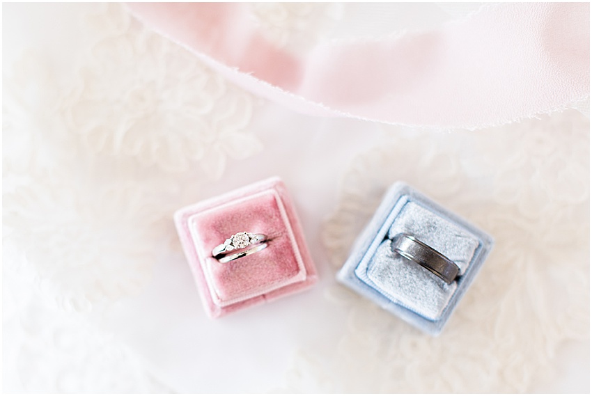 Dusty rose square Velvet ring box with women’s silver wedding band and diamond engagement ring next a baby blue square velvet ring box with a dark grey men’s wedding band 