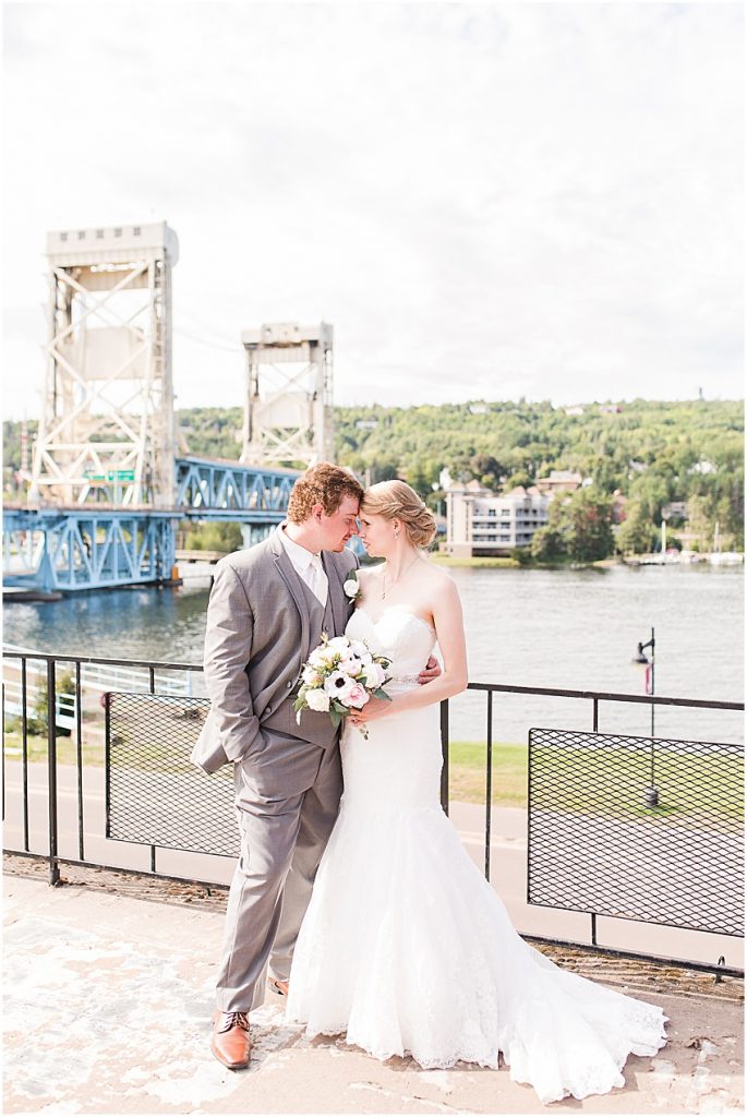 Wedding couple posed forehead to forehead in front of Houghton Michigan’s portage lake lift bridge. Groom is wearing light grey suit and light brown shoes, bride is wearing beautiful mermaid style white gown with lace detailing, no sleeves, and sweetheart neckline. 